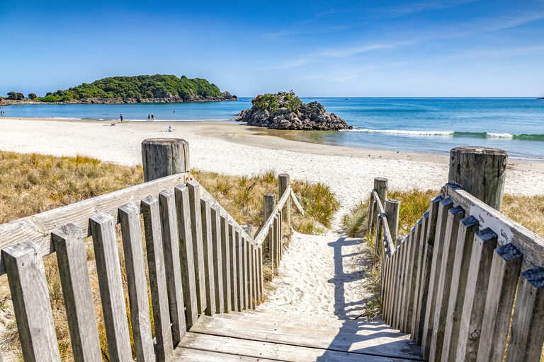 Mount Maunganui, Bay of Plenty, New Zealand - Steps down to beach on a beautiful summer day, with a small island and people walking at the edge of the sea. | Tasman Holiday Parks Waihi Beach