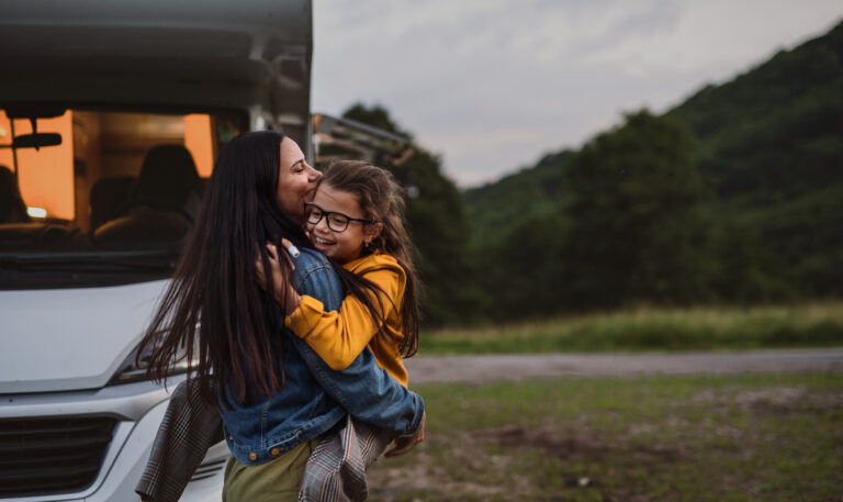 Happy mother with small daughter hugging by car outdoors in campsite at dusk, caravan family holiday trip.