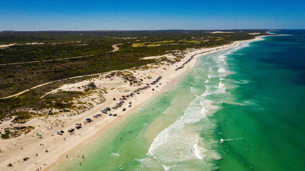 Aerial view of a vast beach with turquoise waters and white sand dunes at Ledge Point. Numerous cars and people are visible along the shoreline, enjoying the bright sunny day. The coastline stretches into the distance, bordered by greenery and a road parallel to the beach.