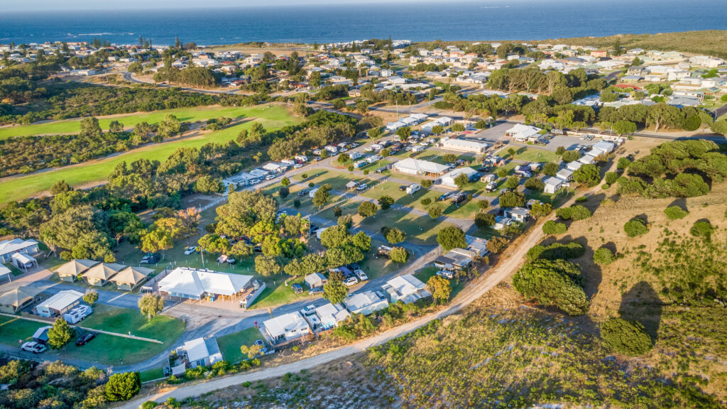 Aerial view of a seaside town at Ledge Point with green spaces and numerous buildings, including houses and mobile homes. Roads wind through the area, connecting homes and green fields. The coastline and ocean are visible in the background, providing a scenic backdrop.