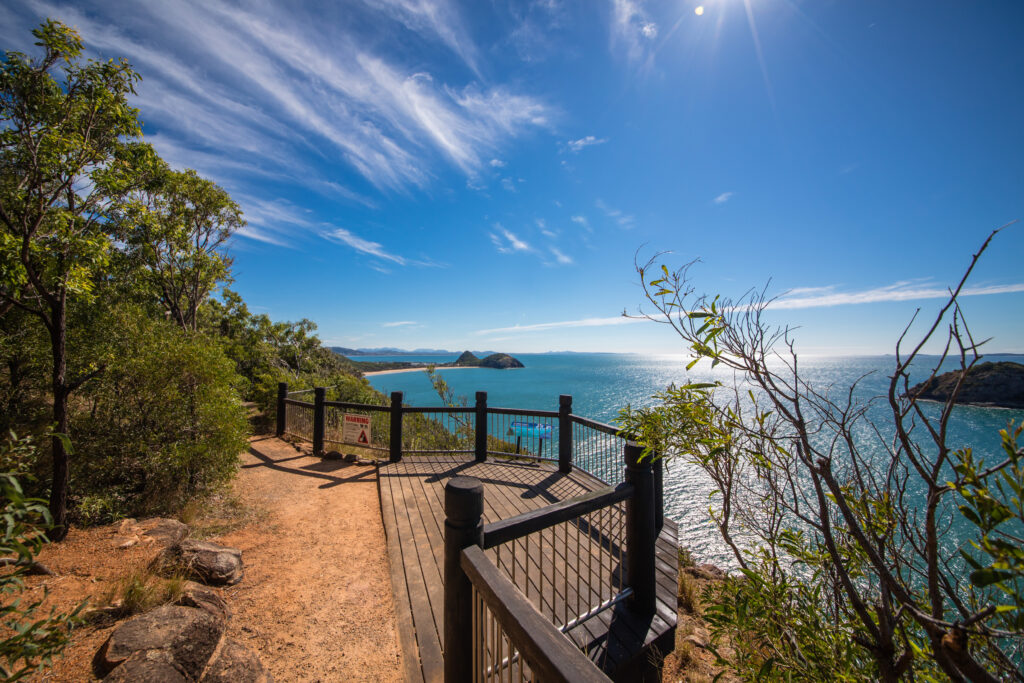 A scenic lookout point with a wooden platform and railing overlooks Yeppoon’s vast, sparkling blue ocean. The sky is clear with a few wispy clouds and the sun shining brightly. Shrubs and trees surround the pathway leading to the platform, with distant land masses visible on the horizon.