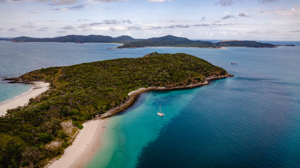 Aerial view of a lush, green island near Yeppoon, surrounded by clear, turquoise waters. The island features sandy beaches and a few boats anchored offshore. In the background, more islands and a hilly landscape can be seen under a partly cloudy sky.