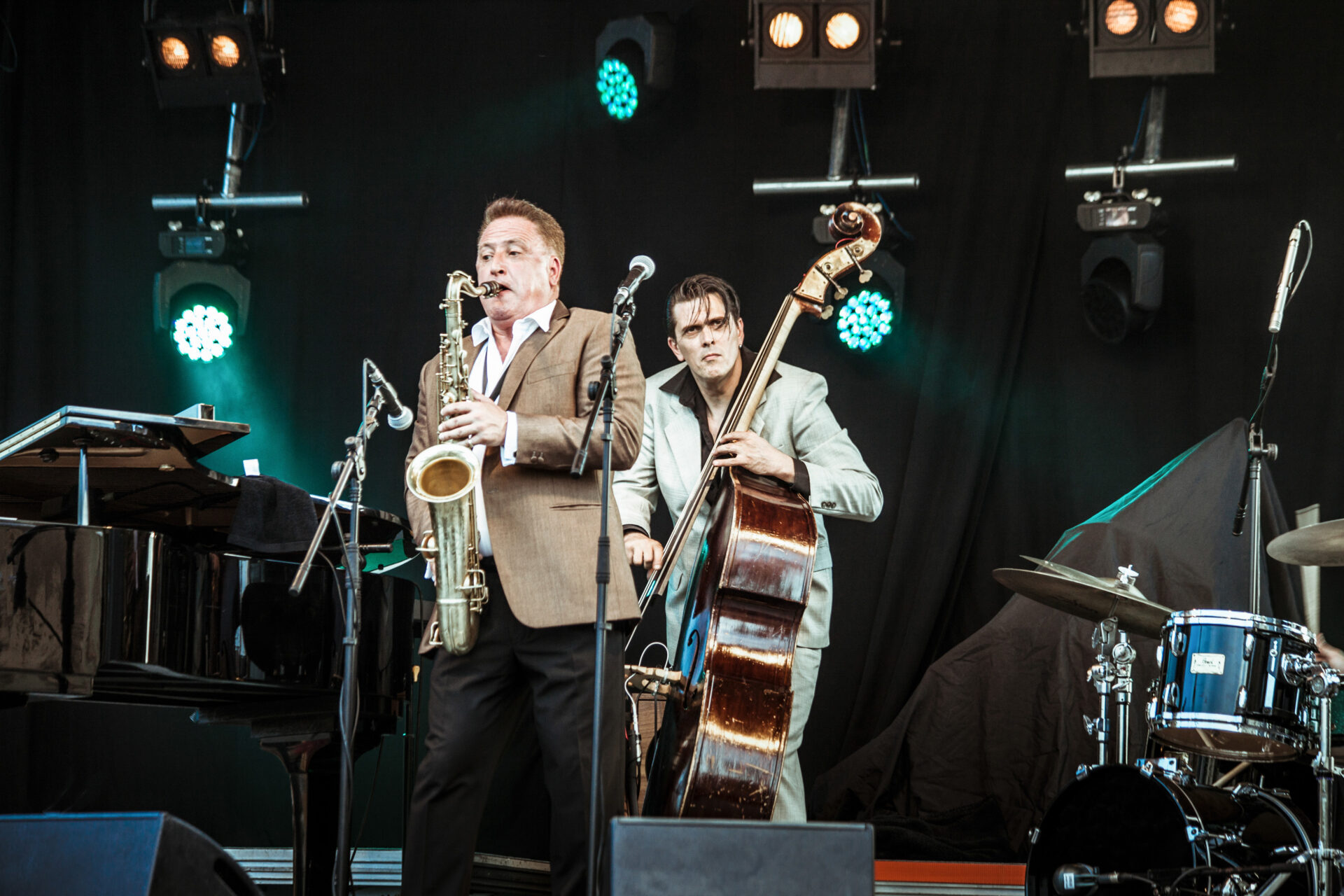 A live jazz performance with three musicians. One plays a saxophone, another a double bass, and the third a piano. They are on a dimly lit stage with spotlights above. The saxophonist and bassist wear suits, and a drum set is visible to the right.