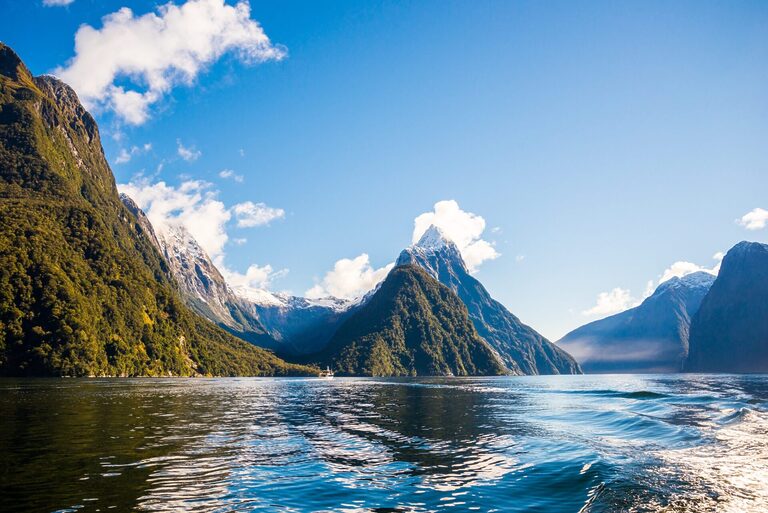 A serene, sunlit scene of a fjord surrounded by steep, lush green mountains with snowy peaks. Calm water in the foreground reflects the blue sky and fluffy white clouds. The scene is tranquil and pristine, with no human presence visible.