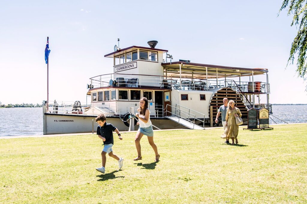 A historic paddle steamer docked by a riverbank under a clear sky. In the foreground, two children are playing and running on the grassy area, while three adults walk nearby. The Australian flag is flying on the steamer, and there is a signboard on the side.