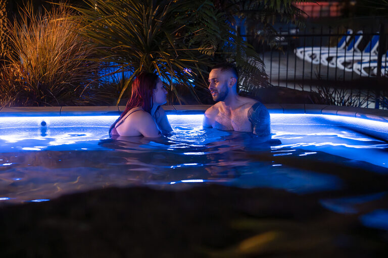 A man and woman are sitting in a pool at night, illuminated by blue lights. They are facing each other and engaged in conversation. The background features lush plants and softly glowing lighting, creating a relaxed, intimate atmosphere.