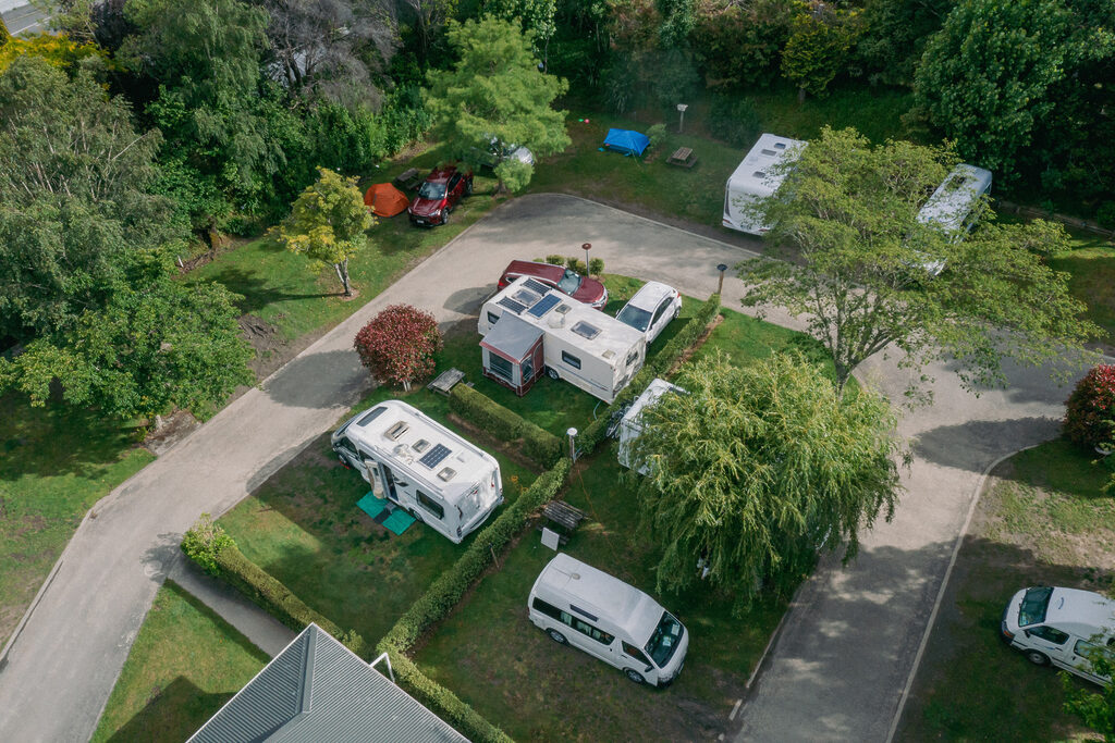 An aerial view of a campground with several RVs parked in designated spots. Green trees and shrubs surround the area, providing a lush backdrop. Tents and camping equipment are visible, alongside access roads and pathways. The scene is bright and peaceful.