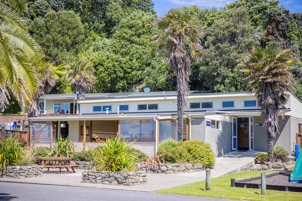 A single-story building with large windows and a flat roof is surrounded by lush greenery and tall palm trees. The entrance features a covered seating area with picnic tables and decorative stone borders. The sky is sunny and clear.