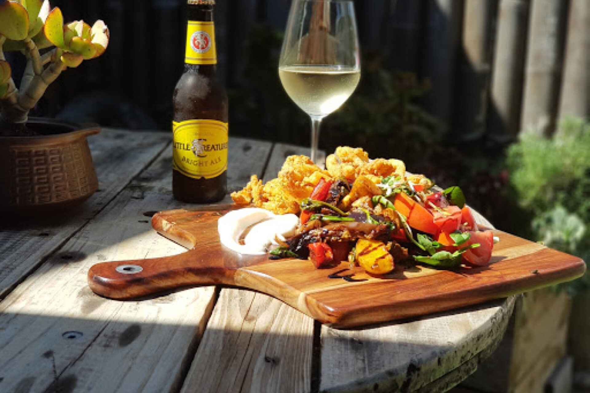 A wooden table holds a small wooden board topped with fried calamari, a colorful tomato salad, and a side of dipping sauce. Next to the board, there's a bottle of amber ale and a glass of white wine. A potted succulent sits to the left, and the table is in a sunny outdoor setting.