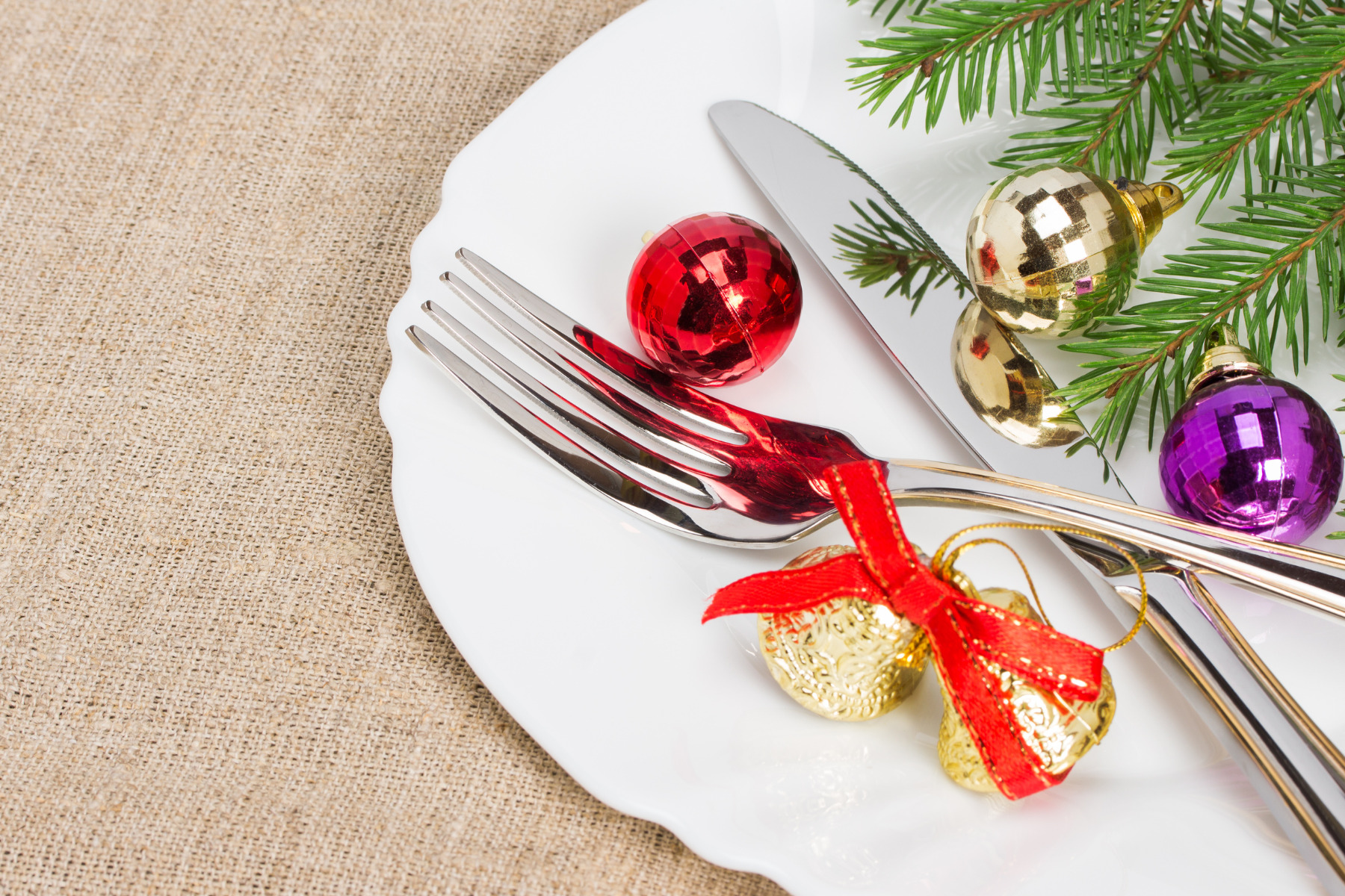 A white plate with silverware and a sprig of evergreen adorned with colorful Christmas ornaments. Two gold jingle bells wrapped with a red bow are placed next to the cutlery. The arrangement rests on a burlap tablecloth, creating a festive holiday setting.