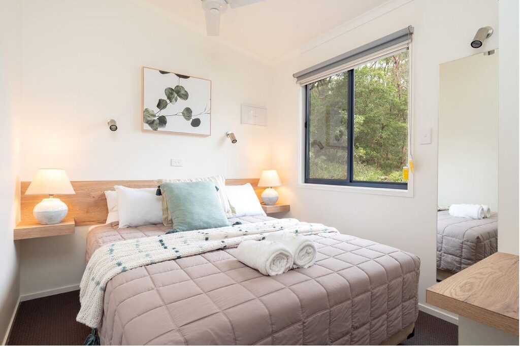 A cozy bedroom with a neatly made bed featuring a beige quilt, pillows, and a folded throw blanket at the foot. Two bedside tables with lamps, a large window with a view of greenery, a framed plant artwork above the bed, and a wall-mounted mirror are also visible.