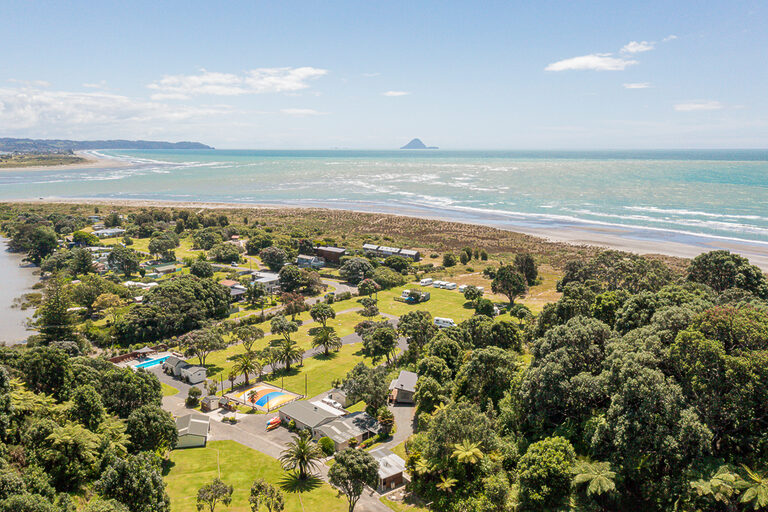 Aerial view of a coastal area with lush greenery, houses, and a campground. The ocean lies beyond a sandy beach, with an island visible in the distance. The scene includes a pool and recreational areas surrounded by trees. The sky is partly cloudy. A great shot celebrating spring in Ohiwa.