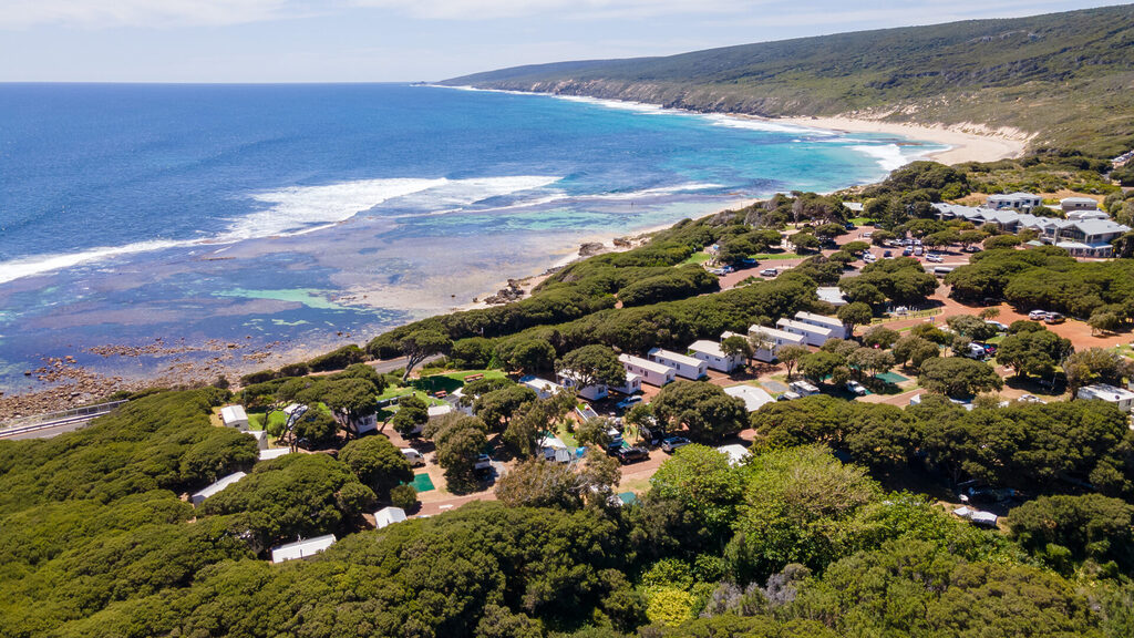 A scenic coastal view featuring a lush green landscape with numerous houses surrounded by trees. The area transitions into a rocky shoreline with clear, turquoise waters extending to the horizon, and a sandy beach is visible in the distance.