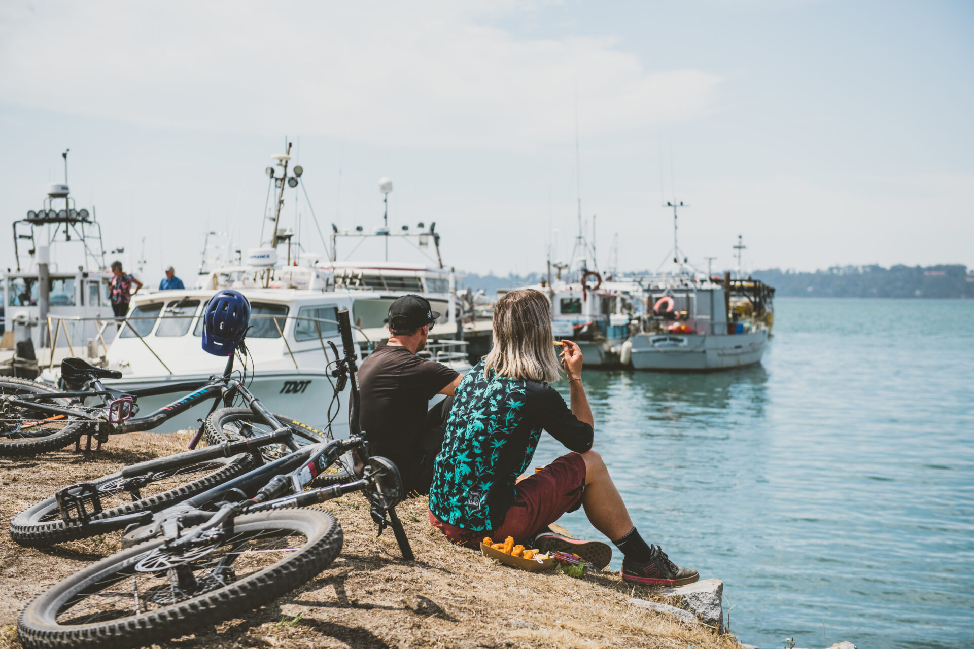 Two people sit on a grassy bank overlooking a marina with several docked boats. Two bicycles are laid on the ground nearby, and one person is eating an orange. The weather is clear, with a blue sky and calm water.