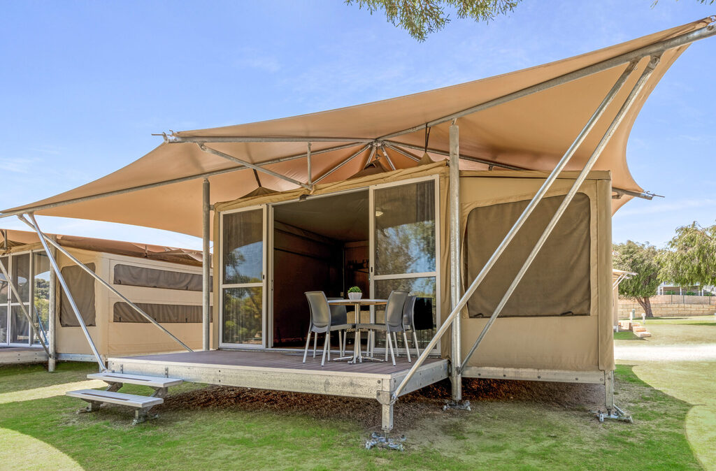 A modern glamping tent with a pitched canvas roof sits on a wooden deck supported by metal frames. The open front has glass doors and windows with beige fabric panels. The deck has a table and chairs, and the tent is on grassy terrain under a clear blue sky.