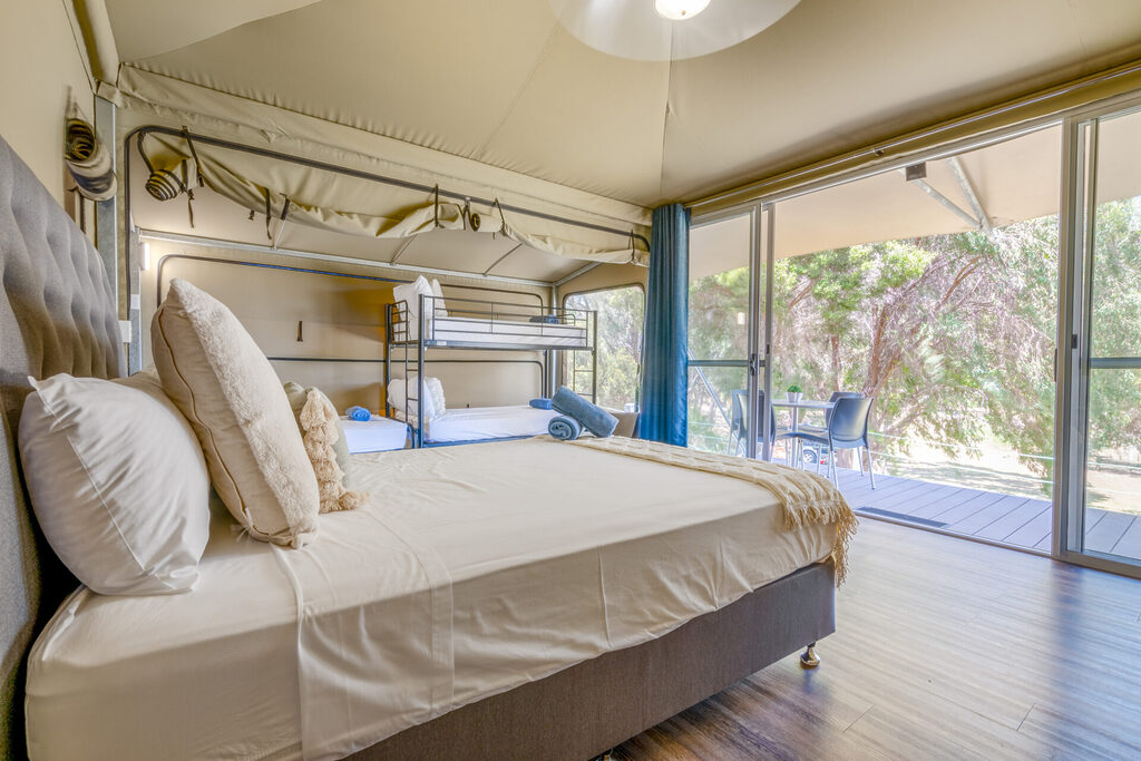 A cozy bedroom features a large bed with white linens and beige pillows, along with bunk beds to the side. The room has wooden flooring and opens to a balcony with a table and chairs, offering a view of nature outside through large sliding glass doors.