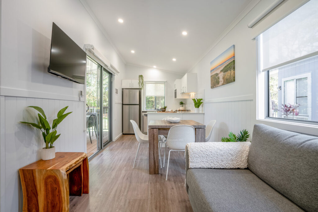 A modern, bright tiny house interior featuring a living area with a gray couch, a wooden table with chairs, a wall-mounted TV, and green plants decor. The kitchen in the back includes a stainless-steel fridge and white cabinets. Sliding glass doors show a deck outside.