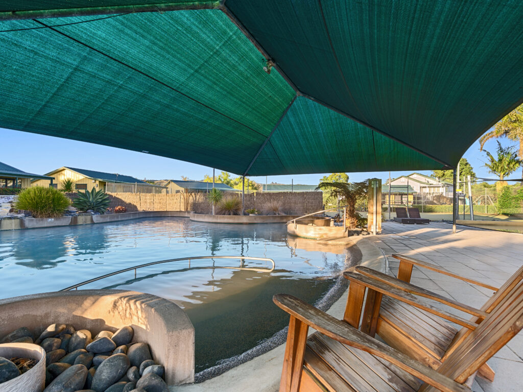 A backyard pool area with a unique design including a rounded shallow end and a submerged ramp. The pool is partially covered by a large green shade canopy. A wooden deck chair is in the foreground, and the surrounding area features plants and a fence.