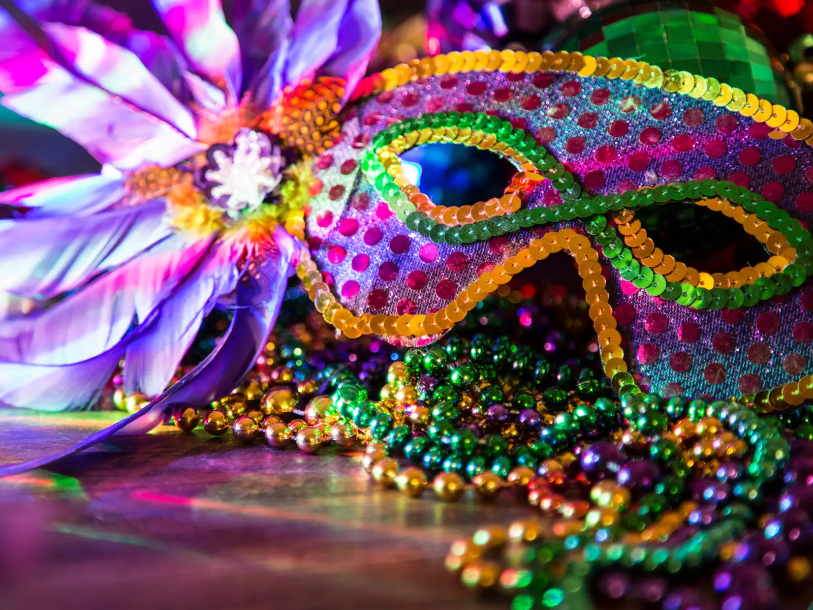 Close-up of a vibrant, sequined Mardi Gras mask surrounded by colorful beads and feathers. The mask features a green and yellow outline with purple accents and is adorned with decorative elements, creating a festive and celebratory atmosphere.