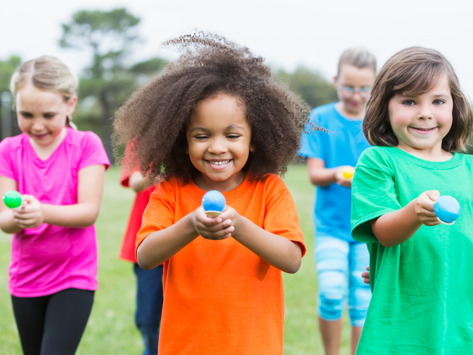 Five children in colorful shirts are participating in an outdoor activity. They are walking forward carefully, holding small balls in spoons. The focus is on a girl in an orange shirt with curly hair, smiling as she concentrates on balancing her ball.