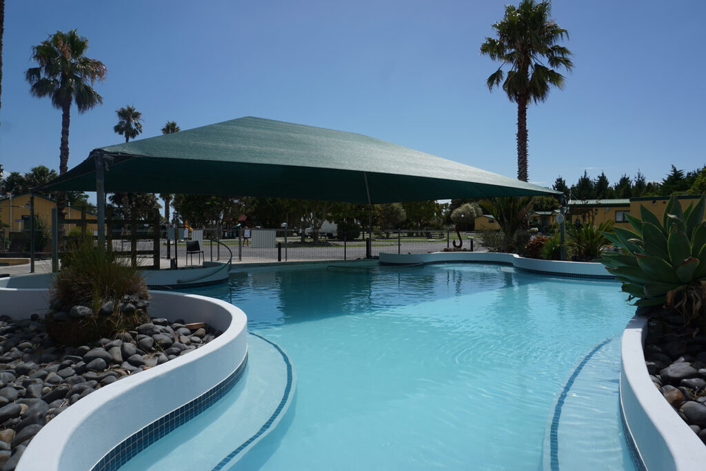 A serene swimming pool filled with clear blue water, surrounded by a sunlit patio and lush greenery.