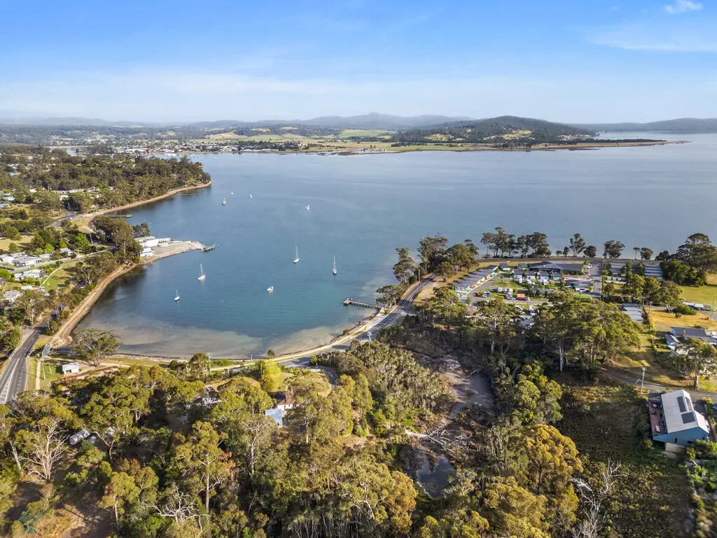 A scenic aerial view of a coastal town featuring a tranquil bay with several anchored sailboats. Surrounding the bay are lush green forests, a few scattered buildings, and a small shoreline road. Hills and mountains are visible in the distant background under a clear blue sky.