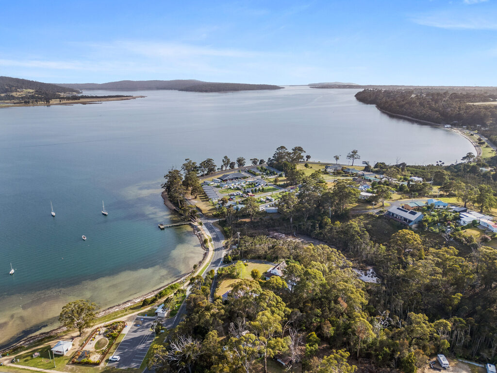 A scenic aerial view of a coastal area featuring a small peninsula with parked boats near the shoreline. The landscape includes lush greenery, buildings, and a winding road adjacent to the water, with a distant horizon of land meeting the blue sky.