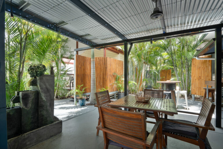 A covered outdoor patio with wooden chairs and a table set surrounded by lush green palm plants. There is a tall water fountain on the left and wooden fencing in the background, creating a tropical, serene atmosphere. String lights decorate the ceiling.