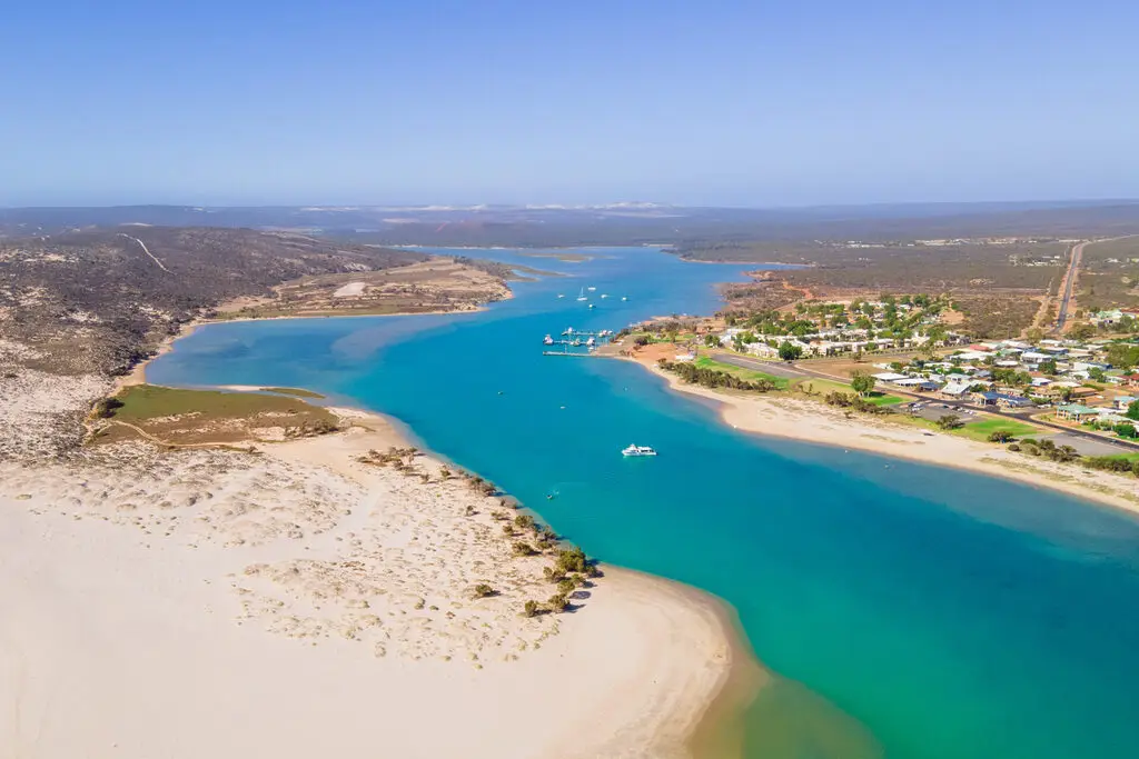 Aerial view of a coastal town featuring a vibrant blue river flowing into a sandy bay. The river is surrounded by dry, hilly terrain on one side and a small settlement with houses and greenery on the other. A few boats are visible on the river. Clear skies above.