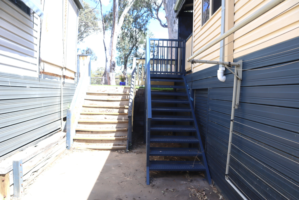 Two metal staircases are positioned between two buildings with siding. On the left, there are wooden steps with metal railings, and on the right, metal steps lead upward with a railing on one side. Trees and sunlight are visible in the background.