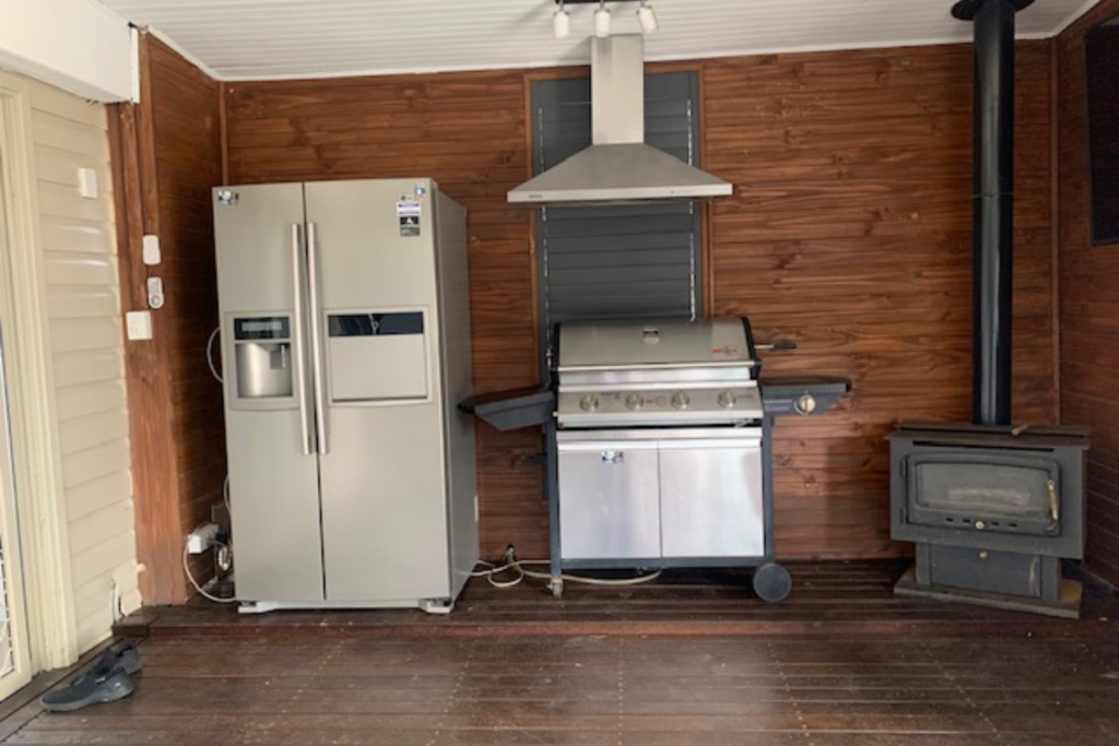 An outdoor kitchen with wooden walls and flooring. It features a stainless steel side-by-side refrigerator, a hooded gas grill in the center, and a wood-burning stove on the right. .