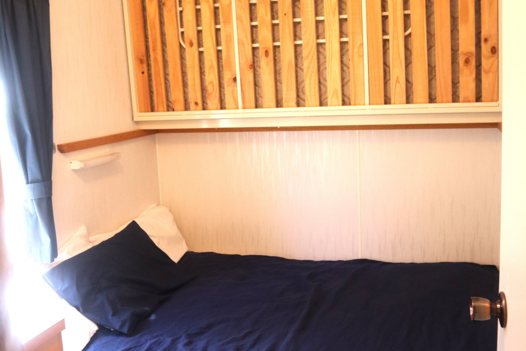 A compact bedroom with a single bed covered in dark blue sheets and white pillows, against a white wall. Above the bed, there are wooden slats arranged vertically, possibly part of a decoration or storage unit. A window with blue curtains is partially visible.