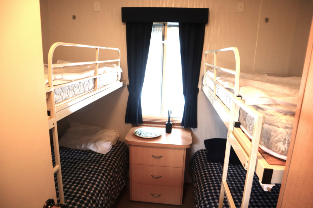 A small bedroom with two bunk beds facing each other. The wooden nightstand in between has a plate and a vase with sticks. A window with closed dark curtains is above the nightstand, letting some light in. The beds are made with checkered blue and white sheets.