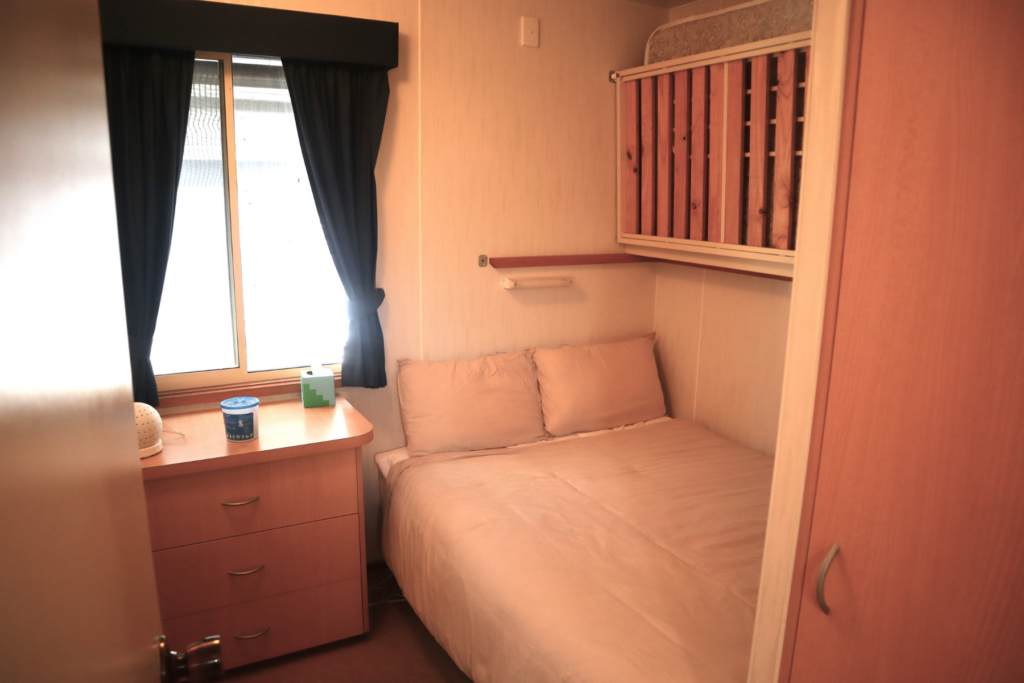 A small, cozy bedroom featuring a bed with white linens tucked against the wall, a window with dark curtains, a wooden nightstand with a few items including a lamp and a tissue box, and a bunk bed overhead with wooden slats. The room has light-colored walls and furniture.