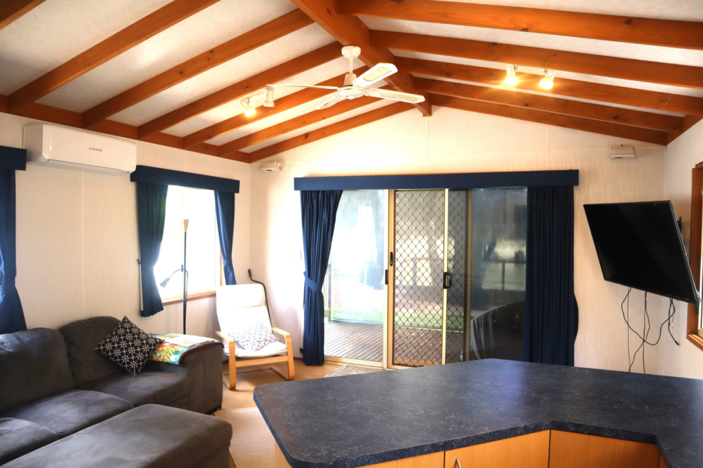 A cozy living room with wooden ceiling beams, a ceiling fan, and a sliding glass door with curtains. The room features a grey sectional sofa, armchair, wall-mounted TV, and a black countertop. Sunlight streams through the window, illuminating the space.