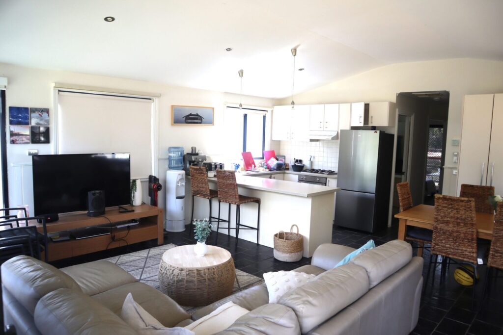 A modern open-plan living area featuring a kitchen, a dining space, and a lounge. The kitchen has white cabinets, stainless steel appliances, and a breakfast bar with stools. The lounge has a grey couch facing a large TV. A dining table with wicker chairs is at the right.