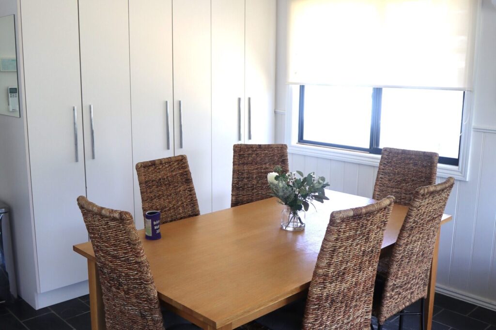 A dining area featuring a wooden rectangular table surrounded by five wicker chairs. A vase with flowers is centered on the table. The room has white-paneled walls, a large window with a roller blind, and white cabinets with silver handles in the background.