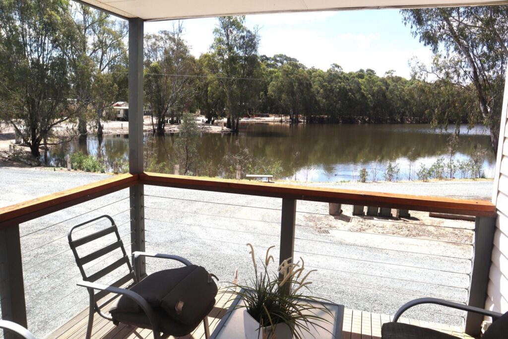 A serene lakeside view from a porch, featuring two chairs and a small table with a plant. The porch is enclosed by a wooden railing with wire lines, overlooking calm water surrounded by trees. The sky is partly cloudy, casting a tranquil atmosphere.