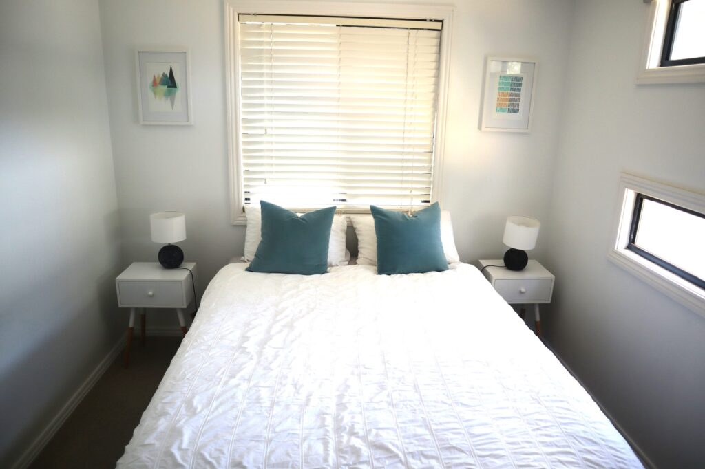 A minimalist bedroom with white walls, a double bed with a white quilt, and two teal cushions. Each side of the bed has a small white nightstand with a lamp. The window above the headboard has closed blinds, and framed artwork hangs on either side.