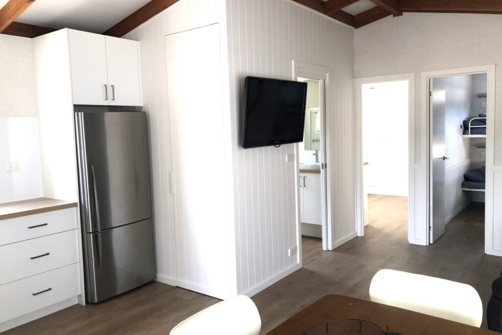 Modern kitchen and hallway. The kitchen features white cabinets, a stainless steel refrigerator, and a wooden countertop. A wall-mounted television is visible. The hallway leads to two open doors, showcasing a bed in the right room. Exposed wooden ceiling beams add warmth.