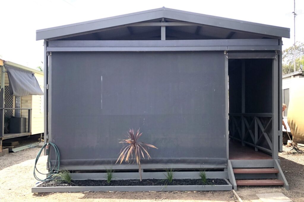 A small, dark gray cabin-like structure with a covered porch area. The front features a screen or curtain, with a single potted plant in front and a row of small plants along the bottom edge. A green garden hose is coiled on the left side.