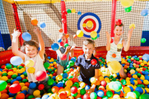 Children are enthusiastically playing in a colorful ball pit, smiling and throwing plastic balls into the air. The ball pit is enclosed with a net, and a target is visible on the back wall. The atmosphere is joyful and lively.