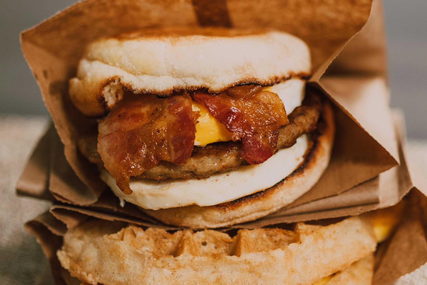 Close-up of a breakfast sandwich with layers of bacon, sausage, egg, and cheese, all nestled between halves of a toasted English muffin. The sandwich is partially wrapped in brown paper.