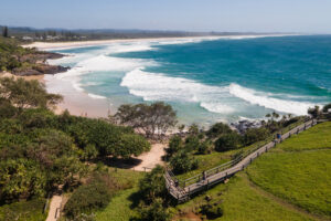 An aerial view of a scenic coastal landscape. The image shows a sandy beach with rolling waves, lush greenery, and a wooden boardwalk leading down to the shore. The background features distant hills under a clear blue sky.