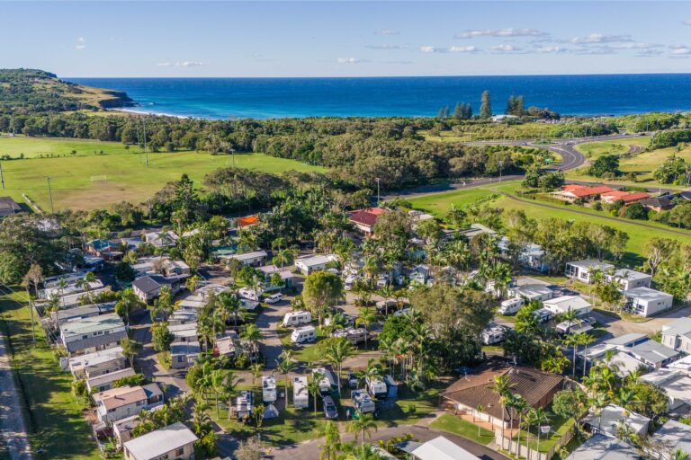 A coastal caravan park in Ballina with numerous RVs and cabins surrounded by lush greenery. A curving road and a few buildings are visible, with the ocean and a distant headland in the background under a clear blue sky.
