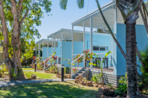 A row of modern, light blue tiny homes or cabins with white roofs and wooden railings are surrounded by lush greenery, palm trees, and vibrant tropical plants in Ballina. A paved walkway winds through the landscaped garden leading to the homes.