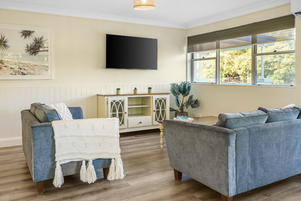 A cozy living room with two blue sofas, a white throw blanket, a TV mounted on the wall, and a patterned sideboard beneath it. The room features large windows allowing natural light, wooden flooring, and a decorative plant in the corner, with a beach-themed picture.