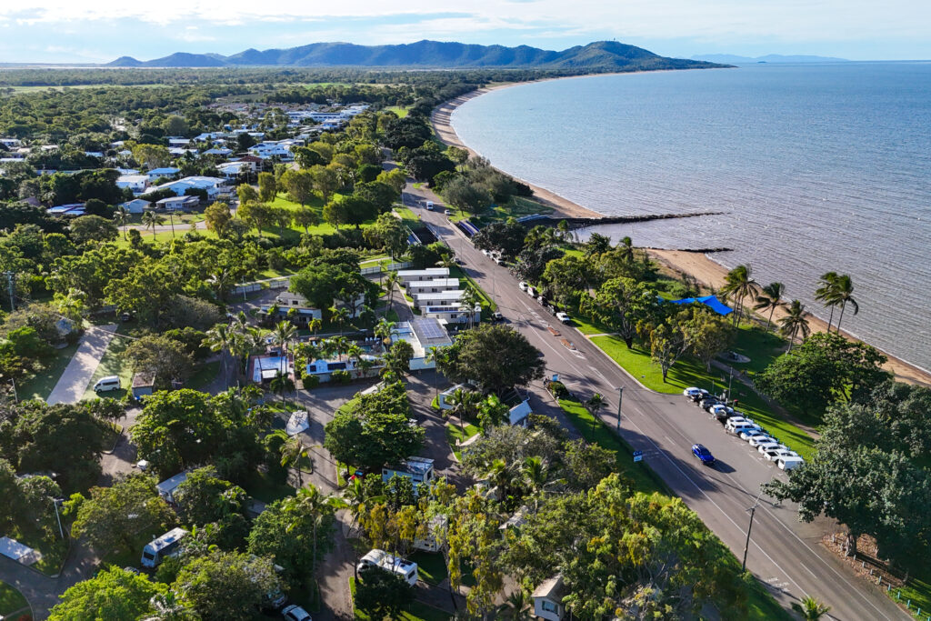 Aerial view of a coastal town with a road running parallel to the beach. There's a mix of residential houses and greenery with mountains in the background and the calm sea stretching out on the right. Several cars are parked along the road.