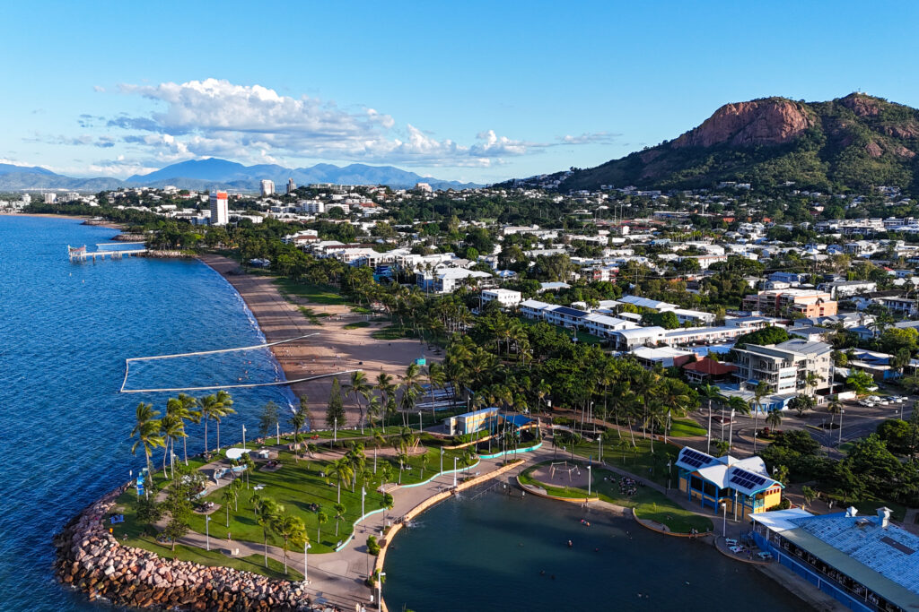 Aerial view of a coastal town featuring a beachfront with palm trees, a public swimming area, and a promenade. Buildings and a rugged hillside are in the background under a clear blue sky.