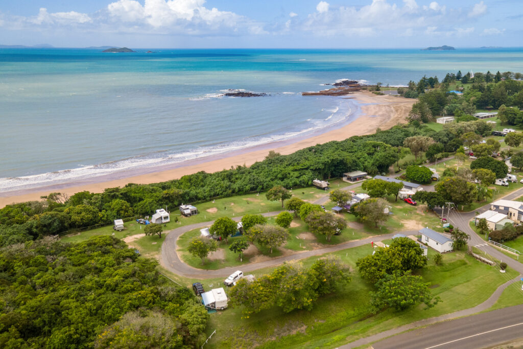 Aerial view of a beachfront holiday park several caravans and motorhomes surrounded by lush greenery. The sandy beach and blue ocean are visible in the background, along with a shoreline of rocks and a few distant islands under a partly cloudy sky.